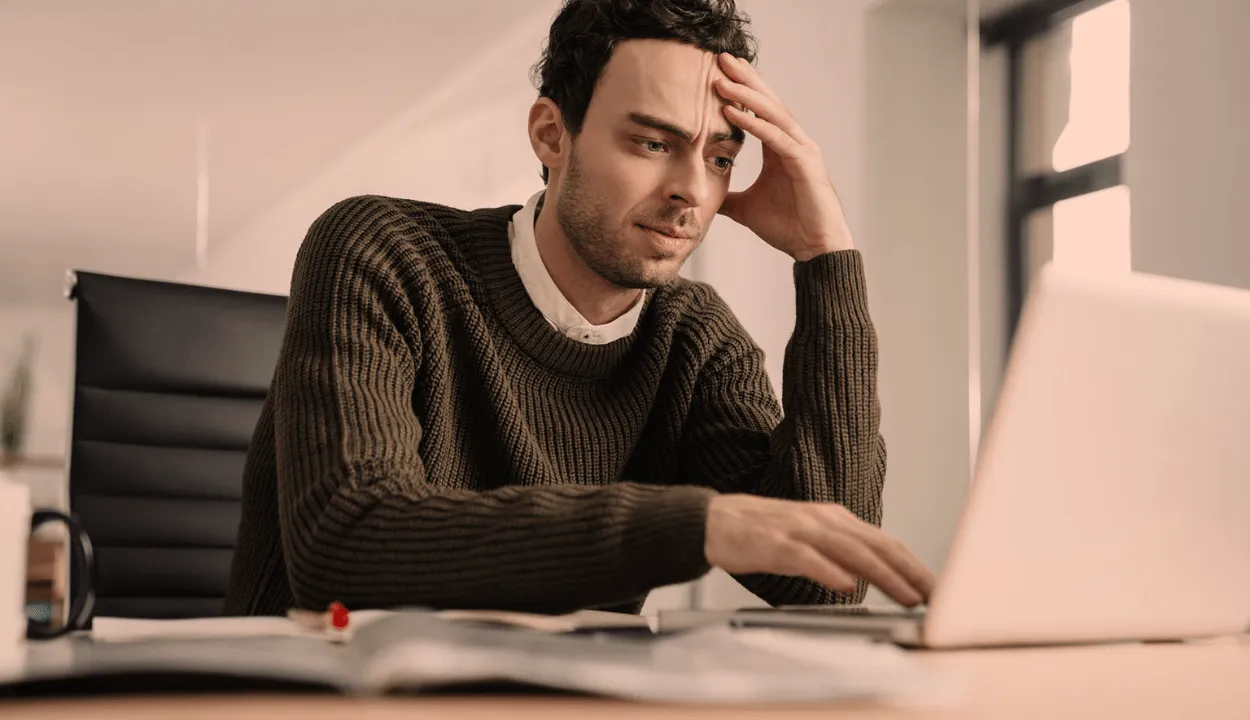 A man sitting in front of a laptop, and one hand is on his forehead, seems like thinking about something