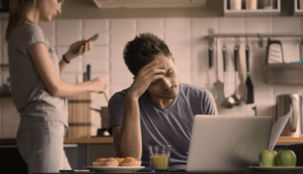 A couple is in the kitchen, the woman is looking at her phone and the man seems upset and looking at his laptop