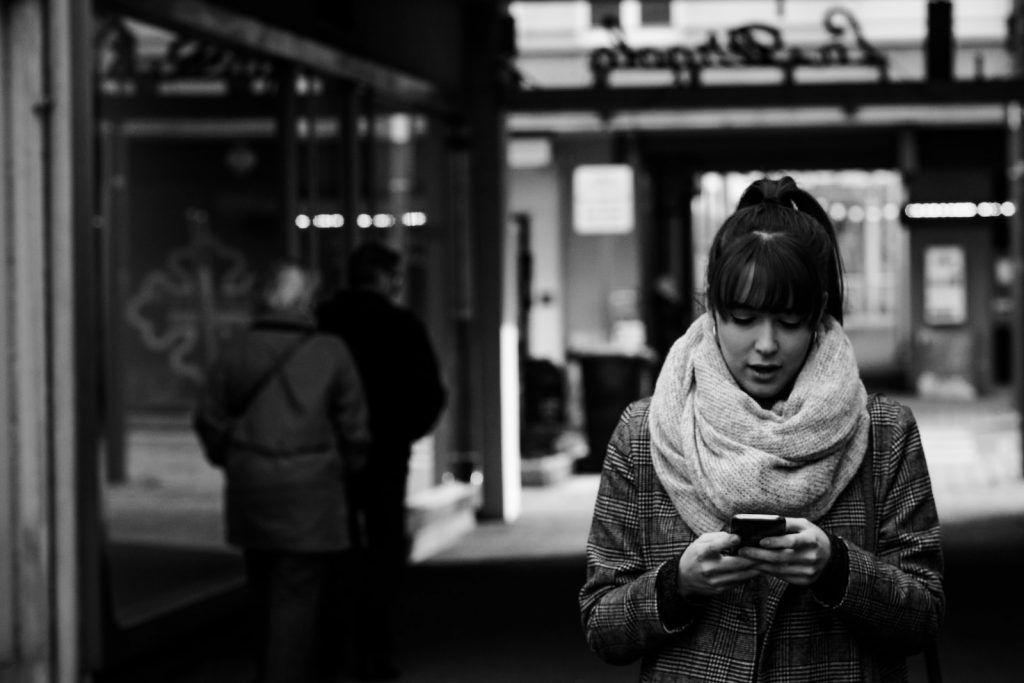 photographie en niveaux de gris d'une femme marchant près de la rue tout en utilisant un smartphone